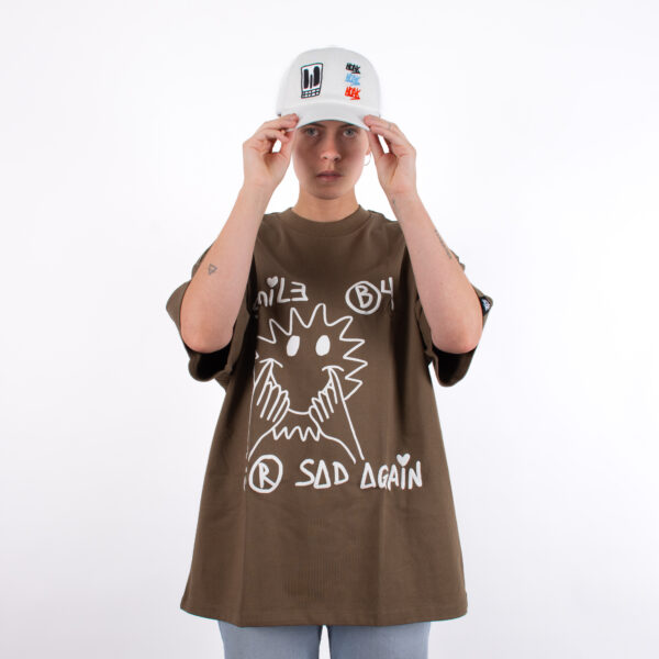 Persona con camiseta marrón de diseño gráfico blanco y gorra blanca con bordados multicolores.