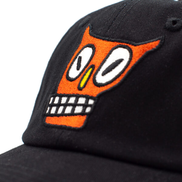 Detalle de bordado en gorra negra con un rostro naranja de ojos grandes y dientes cuadrados blancos.