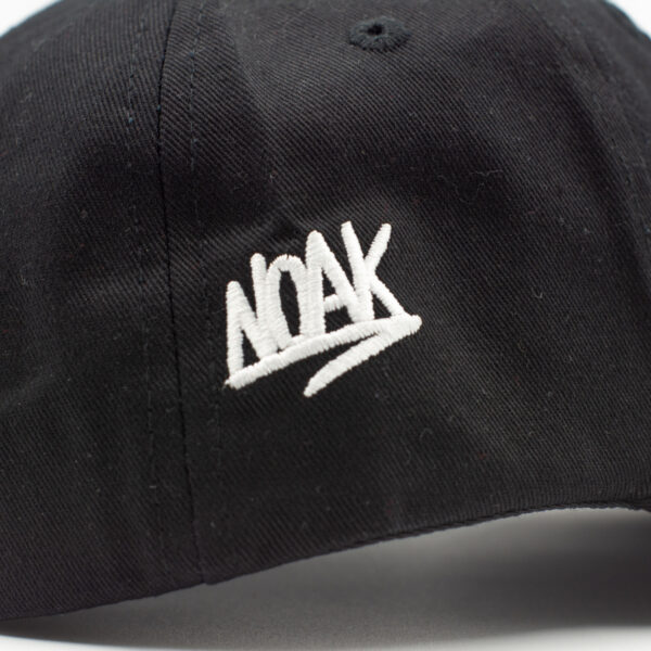 Primer plano de gorra negra con el logo 'NOAK' bordado en blanco sobre tela texturizada.